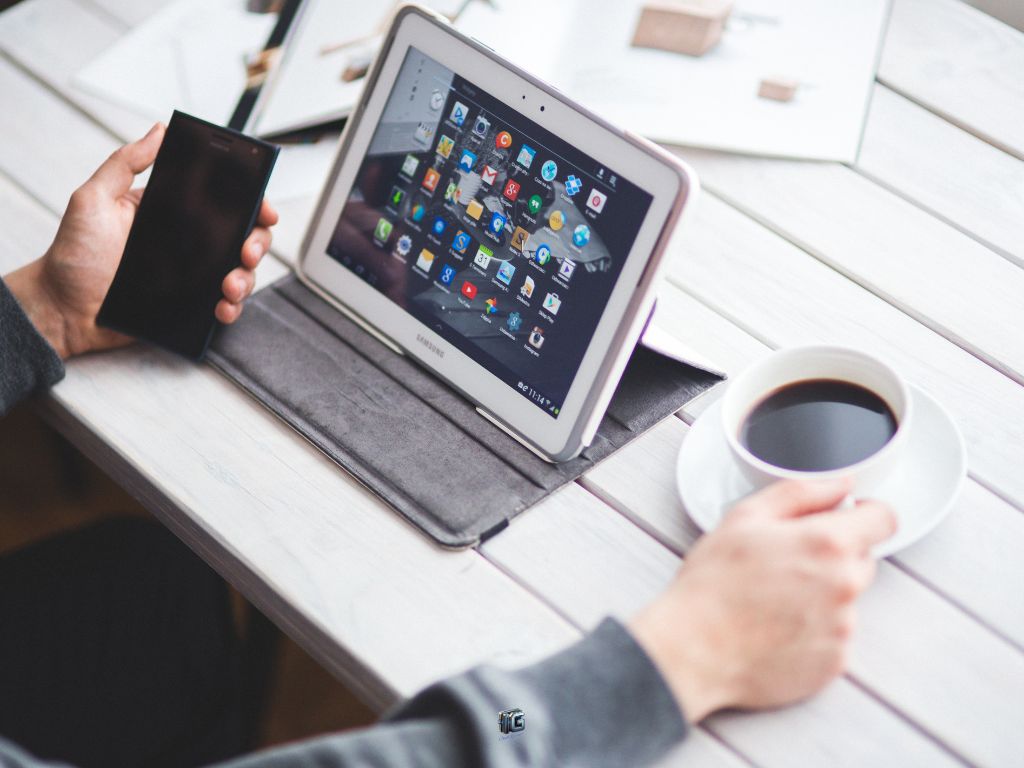 Person using a Samsung tablet with apps on the screen, holding a smartphone and coffee cup at a wooden table.