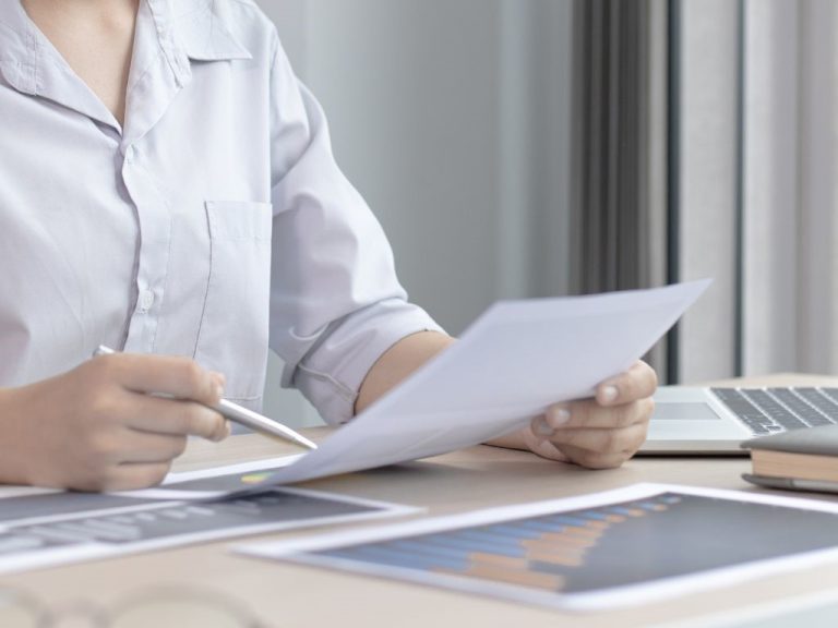 A person holding a pen while analyzing documents at a desk with a laptop.