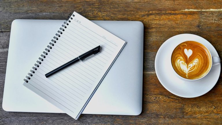 Spiral notebook and black pen on a closed silver laptop, next to a cappuccino with latte art on a wooden table.