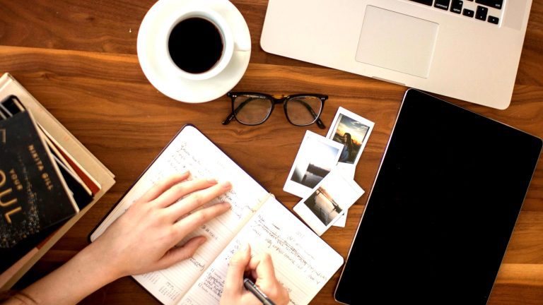 Hands writing a letter in a notebook on a wooden desk with coffee, glasses, and scattered photos.