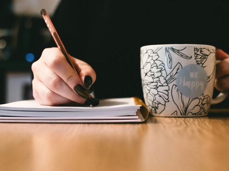 Person writing in a notebook next to a floral mug with 'Be Happy' written on it, symbolizing a calm and inspiring workspace.