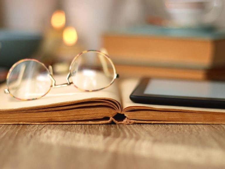 An open book with glasses next to a Kindle, lit warmly.