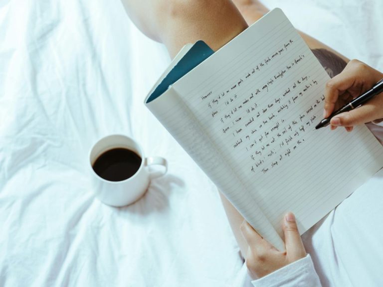 A hand writing in a notebook with a black coffee mug placed on a white bedspread.