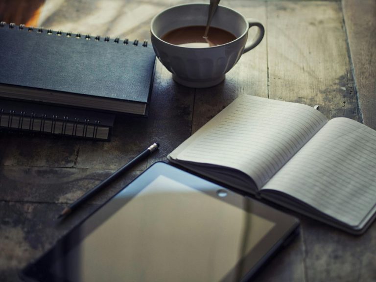 A wooden desk featuring a notebook, a tablet, and a coffee mug with a pencil beside them.