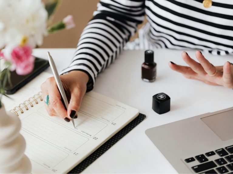 A handwriting in a planner with a laptop and a nail polish bottle on a white desk.