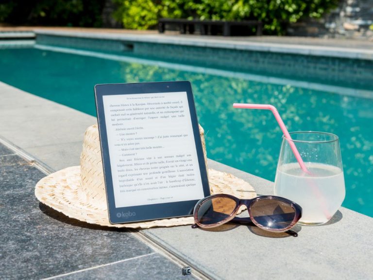 A digital e-reader placed by the poolside on a straw hat, accompanied by sunglasses and a refreshing drink with a straw.