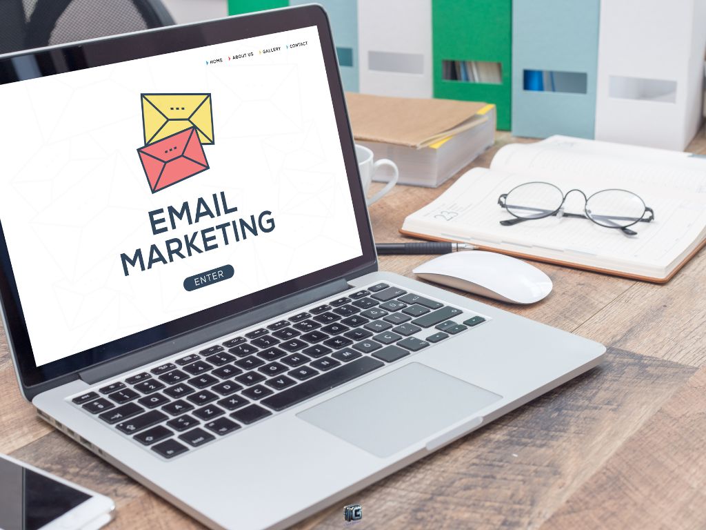 Laptop showing email marketing landing page with colorful envelope icons on a wooden desk with notebook, glasses, and folders in the background.