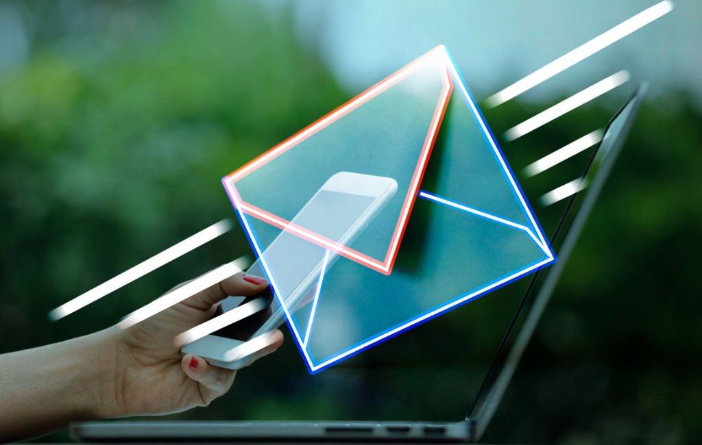 Hand holding smartphone in front of laptop with glowing email icon overlay, symbolizing mobile email marketing and digital communication.