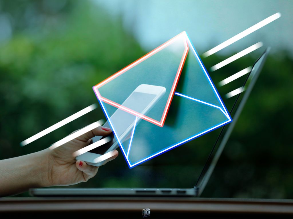 Hand holding smartphone in front of laptop with glowing email icon overlay, symbolizing mobile email marketing and digital communication.