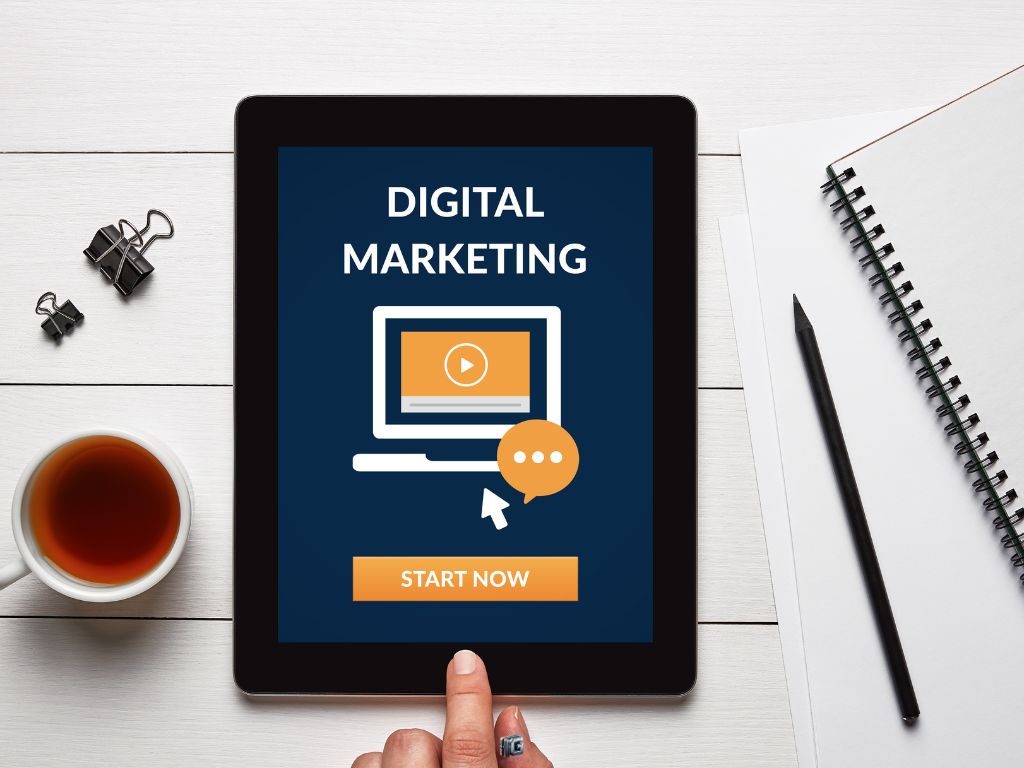 "Tablet on white desk showing digital marketing screen with Start Now button, surrounded by a notebook, pencil, cup of tea, and paper clips."